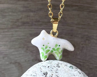 Könnte beinhalten: Eine Halskette mit einem weißen Keramik-Anhänger mit grünem Blattdesign, der an einer goldenen Kette hängt. Der Anhänger hat die Form eines stilisierten Tieres mit vier Beinen.