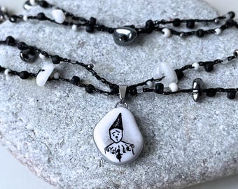 May include: A layered necklace featuring a pendant with a black and white clown design. The necklace has black beaded strands with white and black accents. The pendant is a white stone with a black illustration of a clown.