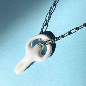 May include: A white ceramic necklace featuring a stylized figure with arms raised above its head. The figure is attached to a silver chain.
