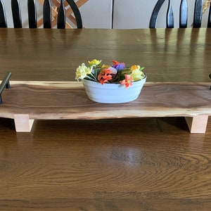 Puede incluir: Una bandeja de servir larga de madera con asas de metal negro. La bandeja está hecha de madera de nogal y tiene un acabado natural de borde vivo. Un pequeño cuenco blanco con flores de colores se encuentra en la bandeja.