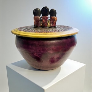 Könnte beinhalten: Eine handgefertigte Schale mit Deckel in tiefem Lila. Die Schale hat einen strukturierten Deckel mit gelbem Rand und drei dunklen, abgerundeten Figuren oben. Die Schale wird auf einem weißen Sockel präsentiert.