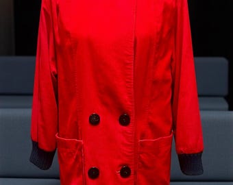 Puede incluir: Chaqueta roja brillante con diseño de doble botonadura. Cuenta con grandes botones negros, dos bolsillos delanteros y puños azul oscuro. El cuello es alto y la chaqueta parece estar hecha de un material suave, posiblemente pana.