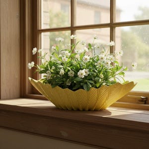 May include: A yellow, leaf-shaped ceramic planter filled with white flowers and green foliage. The planter sits on a wooden windowsill in front of a window with a view of a building.