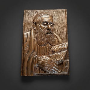 Könnte beinhalten: Eine rechteckige Wandplatte aus Holz mit einem handgeschnitzten Relief einer bärtigen Person, die eine Kopfbedeckung trägt. Die Figur ist mit geschlossenen Augen dargestellt und hält ein Buch oder eine Schriftrolle. Der Hintergrund ist texturiert.