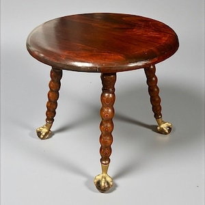 May include: A small, round wooden stool with a dark brown, polished top. The stool has three turned legs with a ball and ring design, and brass-colored claw feet. The wood grain is visible on the seat and legs.