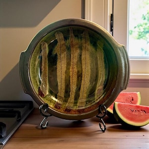 Può includere: Un piatto da portata in ceramica fatto a mano con interno verde e oro, caratterizzato da un design unico e strutturato. Il piatto ha due manici ed è esposto su un supporto in metallo nero. Due fette di anguria sono accanto al piatto.