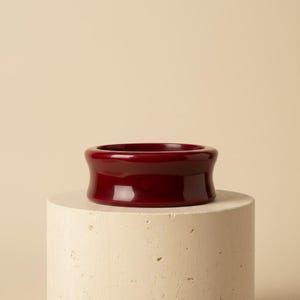 May include: A deep burgundy, wide-band bangle bracelet with a smooth, glossy finish. The bracelet features a concave, curved profile that tapers inward toward the center, creating a distinct hourglass shape around the circumference of the piece.