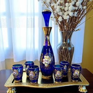 Puede incluir: Un juego de decantador y vasos de chupito azul cobalto con detalles dorados y diseños florales, presentado en una bandeja dorada. Un jarrón alto con flores blancas y un jarrón transparente con tallos secos en el fondo.