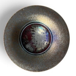 May include: A circular bowl with a wide, textured rim featuring a metallic bronze and gold finish. The deep center of the bowl displays a glossy, dark red base with an iridescent, silver-toned abstract pattern that reflects light with blue hues.