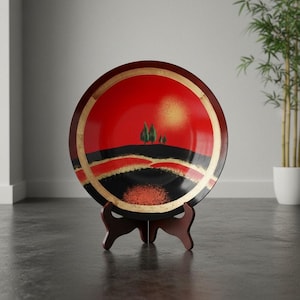 May include: A decorative plate with a red and black landscape design, featuring a golden sun and trees. The plate is displayed on a wooden stand, showcasing its artistic details and vibrant colors.