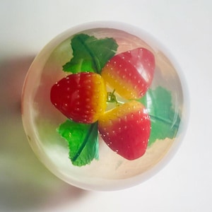 Può includere: Un fermacarte rotondo e trasparente che racchiude tre fragole artificiali e foglie verdi. Le fragole sono rosse e gialle, con una superficie strutturata. Le foglie sono di un verde vibrante. Il fermacarte è posto su uno sfondo bianco.