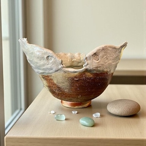 Puede incluir: Un cuenco de cerámica hecho a mano con un diseño único en forma de pez. El cuenco presenta un exterior marrón y beige con una superficie texturizada y un borde de color claro. Pequeñas piedras pulidas en tonos verdes y grises están dispersas alrededor del cuenco.