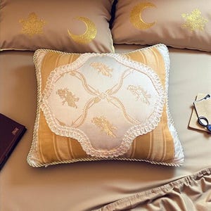 May include: A set of decorative pillows and bedding in a warm, neutral color palette. The pillows feature gold embroidered celestial designs, including moons and stars. One pillow has a striped pattern with a decorative, scalloped edge.