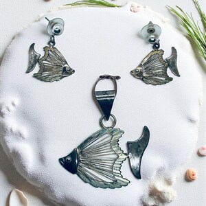 May include: A silver-toned fish pendant and matching earrings set. The earrings are stud style. The fish design features detailed fins and a dark eye. The jewelry is displayed on a white, textured surface with seashells.