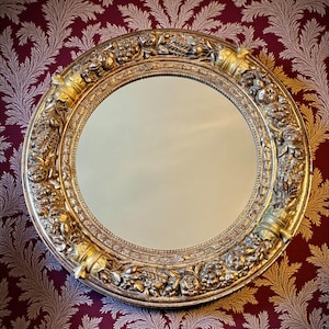 May include: A round, ornate gold mirror with a detailed floral and scrollwork frame. The mirror's frame has a metallic finish and is set against a burgundy wallpaper with a repeating pattern.