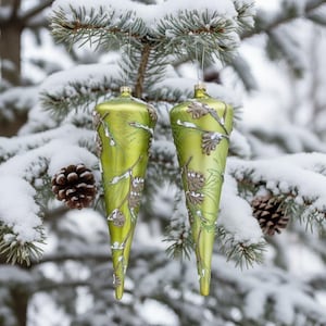 Op de afbeelding: Twee limoengroene kerstversieringen met een druppelvorm, versierd met dennenappel- en takontwerpen. De ornamenten hangen aan zilveren touwtjes tegen een besneeuwde achtergrond van groenblijvende bomen. Een dennenappel rust op een besneeuwde tak.