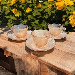 Puede incluir: Tres tazas de té vintage y platillos con un delicado patrón floral. Las tazas y los platillos están hechos de vidrio transparente con un acabado rosa iridiscente. Las tazas están sobre una mesa de madera.