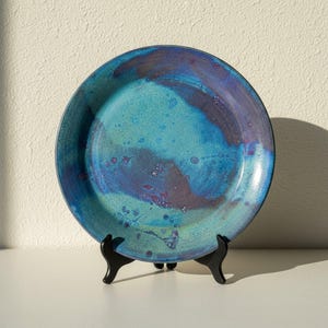 May include: A round ceramic plate featuring a vibrant glaze with swirling patterns of turquoise, deep blue, and hints of purple. The surface displays a mottled, speckled texture throughout, supported by a black plastic display stand.