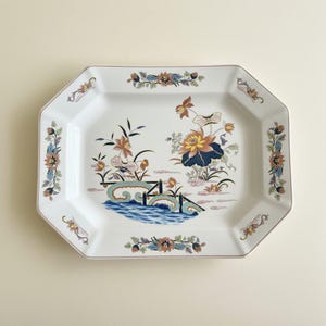 May include: An octagonal white ceramic serving platter featuring a central illustration of a bridge over blue water surrounded by lotus flowers and bamboo stalks. The rim is decorated with repeating floral motifs in orange, blue, and green tones.