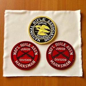 Op de afbeelding: Drie geborduurde patches op een witte doek. De bovenste patch is cirkelvormig met gouden rand, zwarte achtergrond en de tekst "NATIONAL RIFLE ASSOCIATION ANNUAL MEMBER". Twee rode cirkelvormige patches hieronder luiden "NAT'L RIFLE JUNIOR ASS'N DIVISION MARKSMAN."
