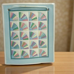 May include: A rectangular ceramic wall hanging with a light blue border and a repeating fan pattern in pastel colors. The fans are arranged in a grid, and a light blue cord is attached at the top for hanging.