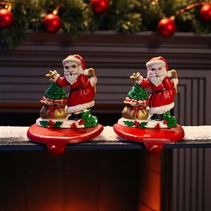 Puede incluir: Dos sujetadores de calcetines navideños con figuras de Papá Noel. Cada Papá Noel viste un traje y gorro rojos, sosteniendo un árbol de Navidad verde y un saco de regalos. Los sujetadores son rojos con un gancho para colgar calcetines.