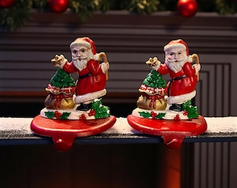 May include: Two Christmas stocking holders featuring Santa Claus figures. Each Santa is dressed in a red suit and hat, holding a green Christmas tree and a sack of gifts. The holders are red with a hook for hanging stockings.