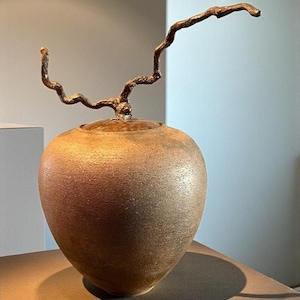 Puede incluir: Un jarrón de cerámica color bronce con una superficie texturizada y una tapa. Un elemento decorativo en forma de rama se extiende desde la parte superior. El jarrón se exhibe sobre una superficie marrón, proyectando una sombra.