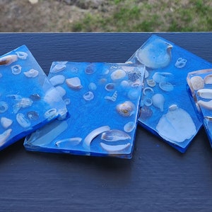 May include: Set of six blue coasters with white seashells embedded in the resin. The coasters are square and have a glossy finish. There is also a matching blue tray with a similar design.
