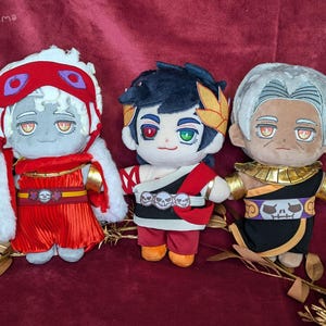 Peut inclure: Trois poupées en peluche de personnages du jeu vidéo Hades. Les poupées ont des traits de visage détaillés et sont habillées de tenues thématiques. La poupée de gauche est rouge et blanche, celle du milieu est rouge, noire et blanche, et celle de droite est noire et dorée.