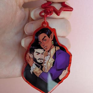May include: Red acrylic keyring with a colourful illustration of two figures embracing. One figure has long black hair, pointed ears, and a purple jacket. The other figure has a beard and is wearing a black suit. The keyring has a red star-shaped top.