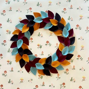 Puede incluir: Una colorida corona de hojas de fieltro. La corona presenta hojas en tonos turquesa, burdeos, negro y dorado. La corona es circular y se muestra sobre un fondo crema con un patrón de hongos repetido.