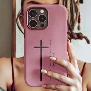 May include: A dusty rose phone case with a black cross and text. The case has a gold-colored camera ring and a black cross design. The text reads "Let all that you do be done in love. 1 Corin 16:14."