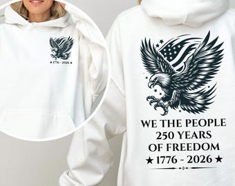 Puede incluir: Sudadera con capucha blanca con bolsillo delantero. Presenta un diseño de águila en blanco y negro y el texto "1776-2026" en la parte delantera. La parte trasera muestra un diseño de águila más grande y el texto "WE THE PEOPLE 250 YEARS OF FREEDOM 1776-2026".