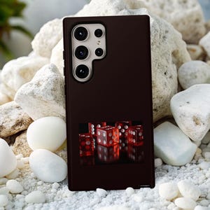 May include: A dark brown phone case featuring a graphic of red dice. The case has a rectangular shape and a camera module with multiple lenses. The background consists of white stones and pebbles.