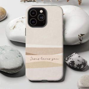 Può includere: Custodia per telefono beige con un design a strati nei toni del beige e del marrone. La custodia presenta il testo "Jesus loves you" in stile scritto a mano. La custodia del telefono ha un modulo fotocamera con tre lenti.