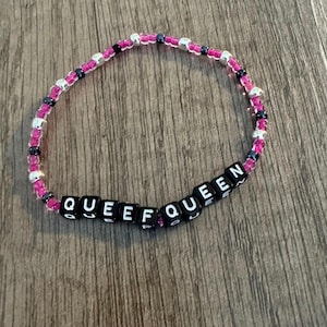May include: A beaded bracelet with the word "QUEEF QUEEN" spelled out in black letter beads. The bracelet features pink, silver, and black beads. The bracelet is on a wooden surface.