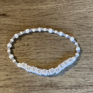 May include: White beaded bracelet with the word "QUEEN" spelled out in white letter beads. The bracelet also features small white and lavender beads. The bracelet is on a wooden surface.