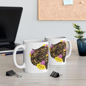 May include: Two white ceramic mugs with a brown and gold snake design, surrounded by pink and yellow flowers. The mugs are on a light-colored surface, with a laptop and office supplies in the background.