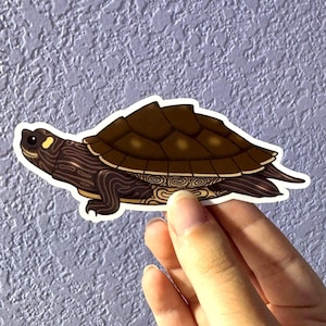 May include: A die-cut sticker featuring a brown turtle with a dark brown, segmented shell and light brown patterned skin. The turtle has a small yellow spot near its eye and is depicted in a side profile view with a white border outline.