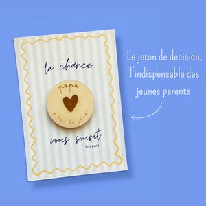 Può includere: Un biglietto di auguri con sfondo a righe blu chiaro e bianco e un disco di legno con un ritaglio a forma di cuore. La carta ha un testo francese che recita "la chance vous sourit (ou pas)" e "papa". Il testo aggiuntivo dice "Le jeton de decision, l'indispensable des jeunes parents".