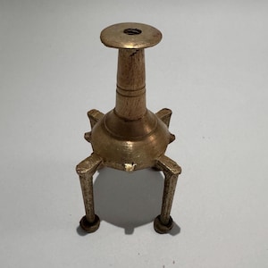 May include: An antique brass candle holder with a round base and three legs. The candle holder has a cylindrical stem and a circular top with a hole for a candle. The brass has a warm, aged patina.