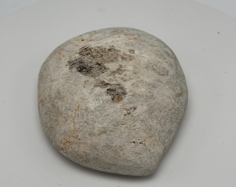 May include: A smooth, gray stone with a rounded shape. The stone has a light color with darker patches and subtle texture variations. The overall appearance is natural and organic, suggesting it could be a decorative element or a natural object.