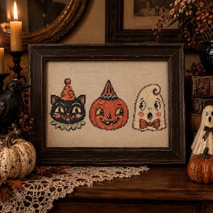 May include: A framed cross-stitch embroidery on light beige fabric featuring three Halloween characters: a black cat wearing an orange party hat, a smiling orange jack-o'-lantern with a striped hat, and a white ghost wearing a brown bow tie.