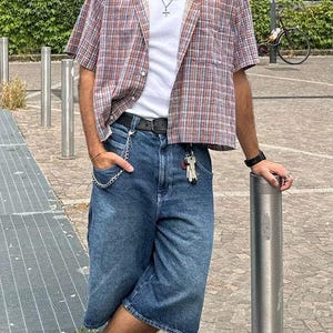 May include: A person wearing a green cap, sunglasses, and a short-sleeved plaid shirt over a white t-shirt. They are wearing blue denim shorts, brown leather loafers, and white socks. A silver chain hangs from the shorts pocket.