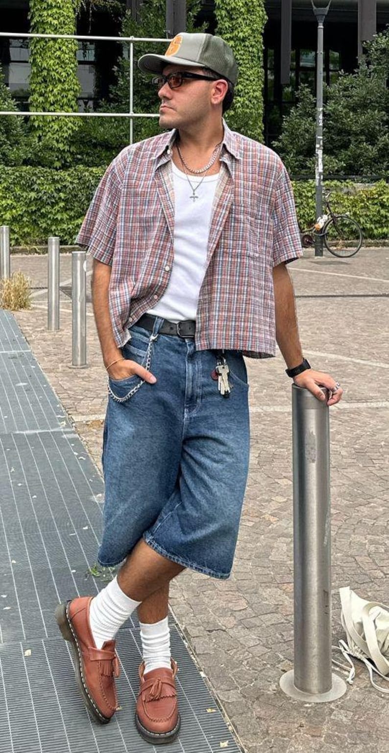 May include: A person wearing a green cap, sunglasses, and a short-sleeved plaid shirt over a white t-shirt. They are wearing blue denim shorts, brown leather loafers, and white socks. A silver chain hangs from the shorts pocket.