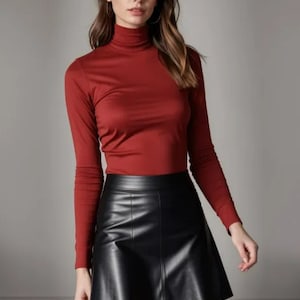 Op de afbeelding: Een vrouw draagt een rode trui met een hoge hals en een zwarte leren rok.