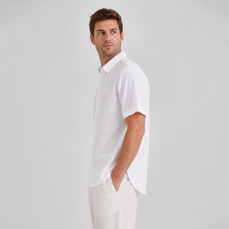 May include: A short-sleeved white button-down shirt with a classic collar and a textured fabric finish. The shirt features a straight hemline and is worn tucked into matching white trousers, showcasing a clean and simple design aesthetic.