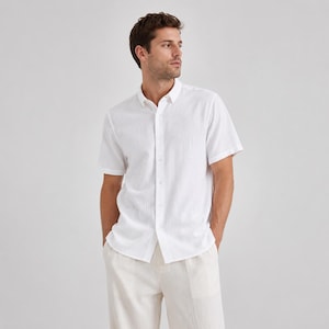 May include: A crisp white short-sleeved button-down shirt with a subtle textured fabric finish, featuring a classic collar, a front button placket, and a straight hemline, paired with light beige trousers in a neutral, clean aesthetic.