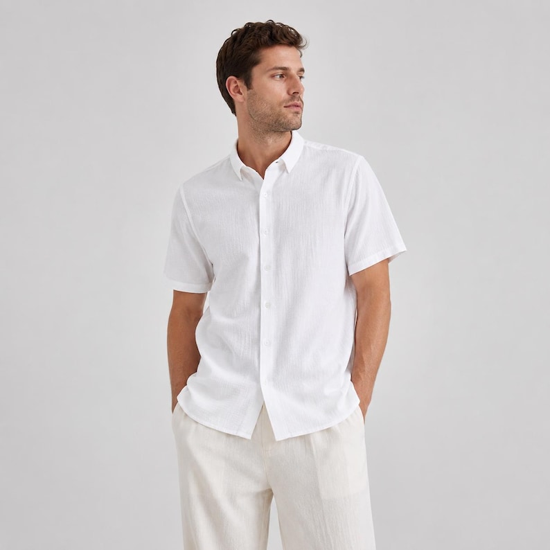 May include: A crisp white short-sleeved button-down shirt with a subtle textured fabric finish, featuring a classic collar, a front button placket, and a straight hemline, paired with light beige trousers in a neutral, clean aesthetic.